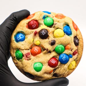 May include: A round, golden-brown baked cookie embedded with numerous colorful M&M chocolate candies in shades of red, blue, green, yellow, and brown. The surface of the cookie has a textured, slightly uneven appearance with visible chocolate pieces.