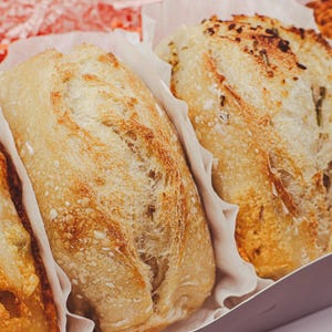 May include: A box of assorted artisan bread loaves. The loaves vary in color and toppings, including a loaf with green herbs and cheese. The bread is presented in a cardboard box with white paper liners, ready for serving.