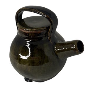 May include: A round, dark brown glazed ceramic teapot with a glossy finish. It features a short, wide spout extending from the side and a curved handle arched over the top. The lid is integrated into the top opening, matching the dark, reflective glaze.