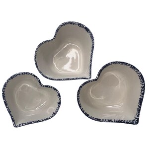 May include: Three heart-shaped ceramic bowls. The bowls are white with a dark blue dotted trim around the edges. The bowls are different sizes and are arranged in a group. The bowls are empty and have a glossy finish.