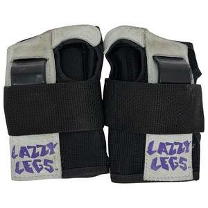May include: Pair of black wrist guards with gray suede-like padding and black straps. Each guard features a rectangular patch with the words "LAZZY LEGS" in purple lettering. The guards are designed for wrist protection during sports or activities.