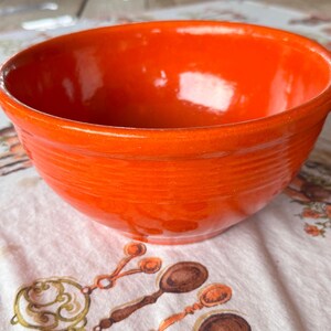 May include: A bright orange ceramic bowl with a smooth, glossy finish and a series of horizontal ridges encircling the exterior. The bowl features a wide, open top and a rounded base, showcasing a vibrant, solid color throughout the entire piece.