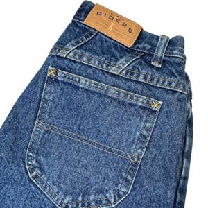 May include: Close-up of blue denim jeans with a back pocket and a brown leather label that reads "DENIM RIDERS". The jeans have yellow stitching and a classic five-pocket design. The denim is a medium wash.