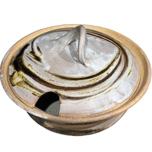 May include: A ceramic lidded serving dish with a handle. The dish is beige with brown and green swirl patterns on the lid. The rim and handle are a lighter beige color. There is a small opening on the side of the dish.