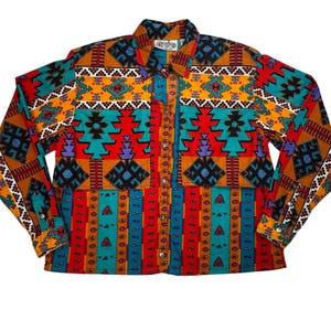 May include: A button-down shirt with a vibrant Southwestern pattern in red, orange, teal, and black. The shirt features a collared neckline, long sleeves, and a Rodeo brand label. The design includes geometric shapes and vertical stripes.