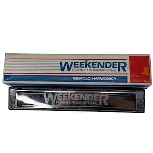 May include: A Hohner International Weekender tremolo harmonica with its original box. The harmonica is silver with the word "Weekender" printed on it. The box is white with blue and red stripes and the word "Weekender" printed on it.