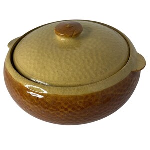 May include: A round ceramic casserole dish with a lid, featuring a textured, dimpled surface. The lid is a light beige color, while the base of the dish is a darker, rich brown. The pot includes two small side handles and a central knob on the lid.