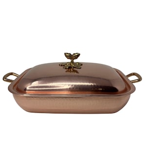 May include: A copper-colored rectangular casserole dish with a lid and handles. The lid is topped with a decorative gold-colored leaf-shaped handle. The dish has a hammered texture and is designed for cooking and serving.
