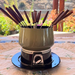 May include: A vintage olive green fondue pot with a black base and a temperature control dial labeled Cornwall. The pot contains a set of metal fondue forks with dark wood handles, each topped with a small colored tip in blue, yellow, or red.