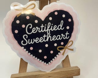 Könnte beinhalten: Ein herzförmiger Aufkleber mit dem Schriftzug "Certified Sweetheart" in weißer Schrift. Der Aufkleber hat einen schwarzen Hintergrund mit weißen Punkten, einen rosa gewellten Rand und goldene Schleifenakzente. Er wird auf einer kleinen Holzstaffelei präsentiert.