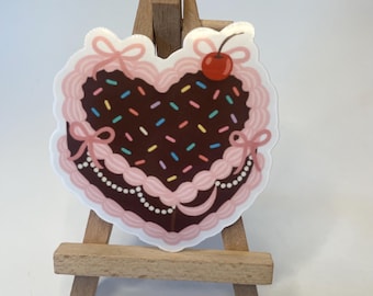 May include: A heart-shaped sticker resembling a chocolate cake with pink frosting, sprinkles, and a cherry on top. The sticker has a white border and is decorated with pink bows and a pearl-like border. It is displayed on a small wooden easel.