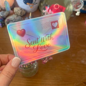 以下が含まれることがあります： 「Sent With Love」の文字が筆記体で書かれた、封筒型のホログラフィックステッカー。赤いハートとハート型の切手が特徴です。ステッカーは虹色の光沢があります。