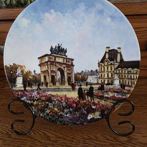 May include: Decorative plate featuring a painted scene of a European city with an archway and buildings. The plate is displayed on a black metal stand. The artwork includes a blue sky and colorful flowers.