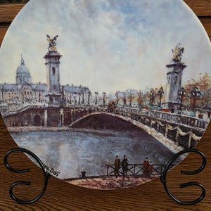 May include: A decorative plate featuring a scenic painting of a bridge over a river, with buildings and statues in the background. The plate is displayed on a black metal stand, showcasing the artwork's details and colors.