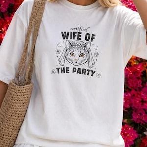 Op de afbeelding: Een wit T-shirt met korte mouwen en een zwarte afbeelding van een kat met een bruidssluier en de tekst "certified WIFE OF THE PARTY", omringd door kleine bloem- en maanpictogrammen. Het shirt wordt gecombineerd met een beige geweven tas.