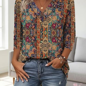 Könnte beinhalten: Eine V-Ausschnitt-Bluse mit einem lebendigen, mehrfarbigen Paisley- und Blumenmuster in Orange-, Petrol-, Magenta- und Goldtönen, kombiniert mit einer hellblauen, abgenutzten Jeans. Die Bluse hat Dreiviertelärmel und eine lockere Passform.