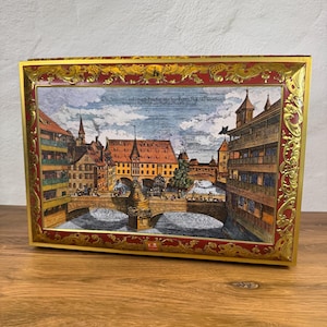 Könnte beinhalten: Eine rechteckige Metalldose mit einer detaillierten historischen Abbildung einer europäischen Stadt mit Steinbrücke, Fluss und Fachwerkhäusern. Die Szene ist von einem verzierten rot-goldenen Rahmen mit kunstvollen Schnörkelmustern umgeben.