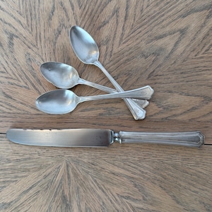 May include: A set of three silver-toned metal spoons and one matching dinner knife. Each piece features a slender handle with a subtle engraved detail at the end. The knife has a long, flat blade with visible dark spots, while the spoons have oval bowls.
