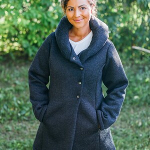 May include: A woman wearing a dark gray, long-sleeved, collared jacket with a shawl collar. The jacket has a single button closure and pockets on the sides. She is standing in front of a green background.
