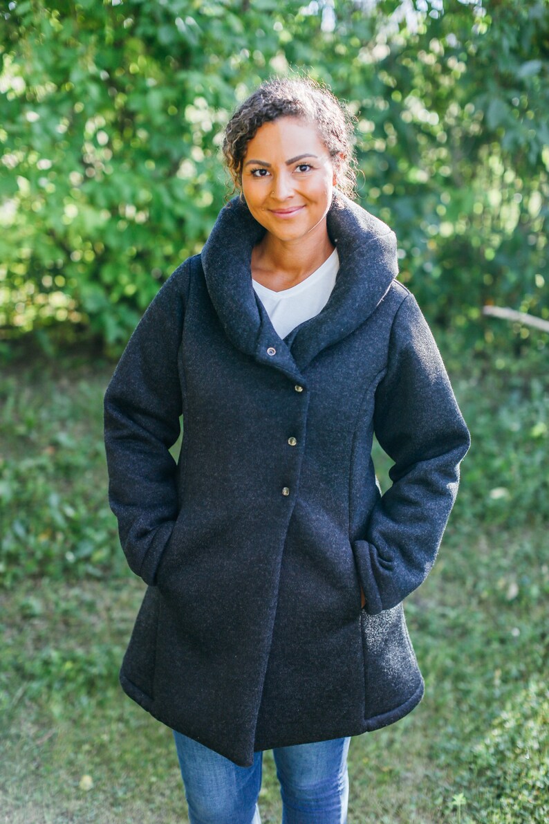 May include: A woman wearing a dark gray, long-sleeved, collared jacket with a shawl collar. The jacket has a single button closure and pockets on the sides. She is standing in front of a green background.