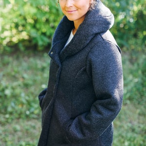 May include: A woman wearing a dark gray hooded coat with a high collar. The coat has a zipper closure and pockets. She is standing in front of a green background.