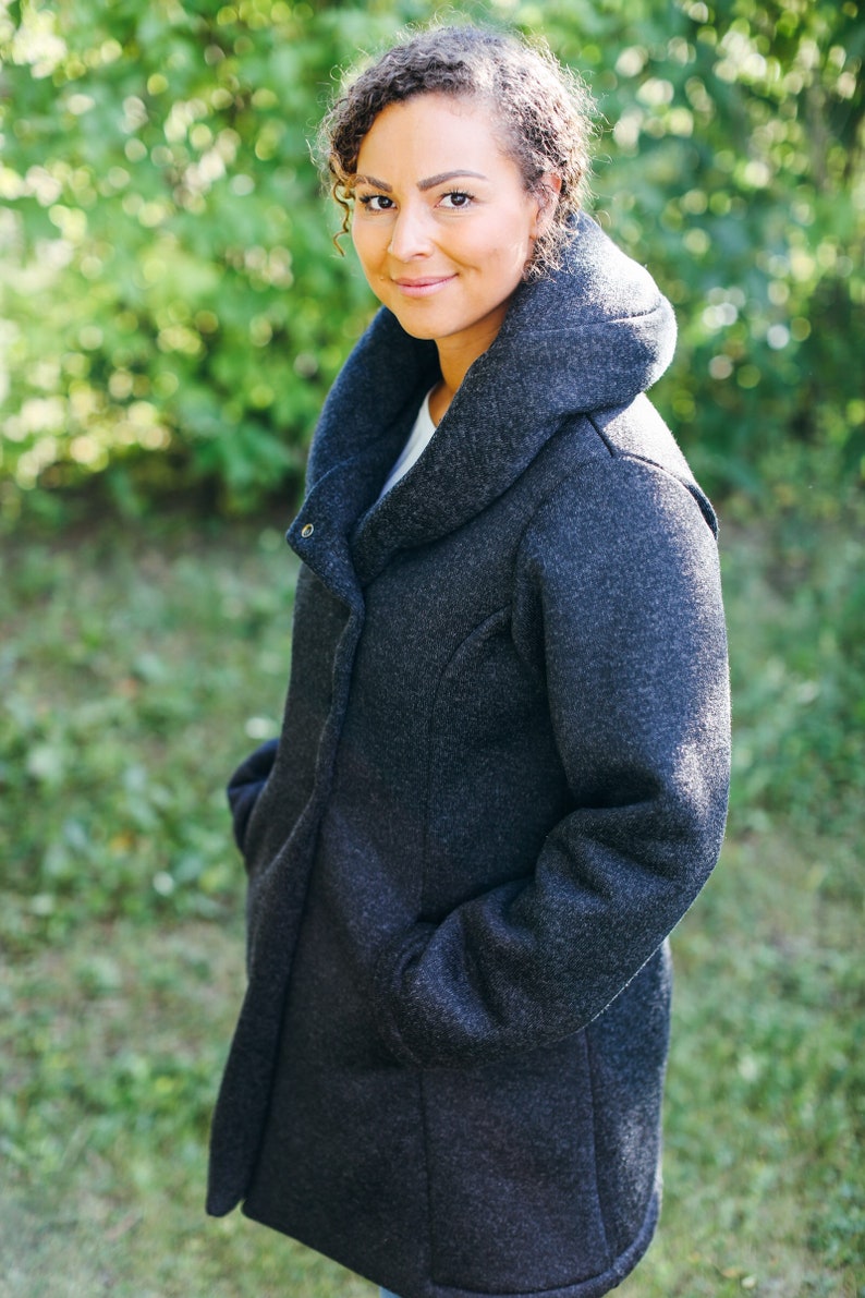 May include: A woman wearing a dark gray hooded coat with a high collar. The coat has a zipper closure and pockets. She is standing in front of a green background.