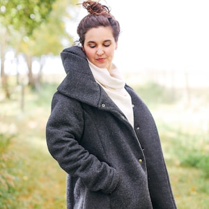 May include: A woman wearing a dark grey, long-sleeved coat with a high collar and a white turtleneck sweater underneath. The coat has a button closure and pockets.