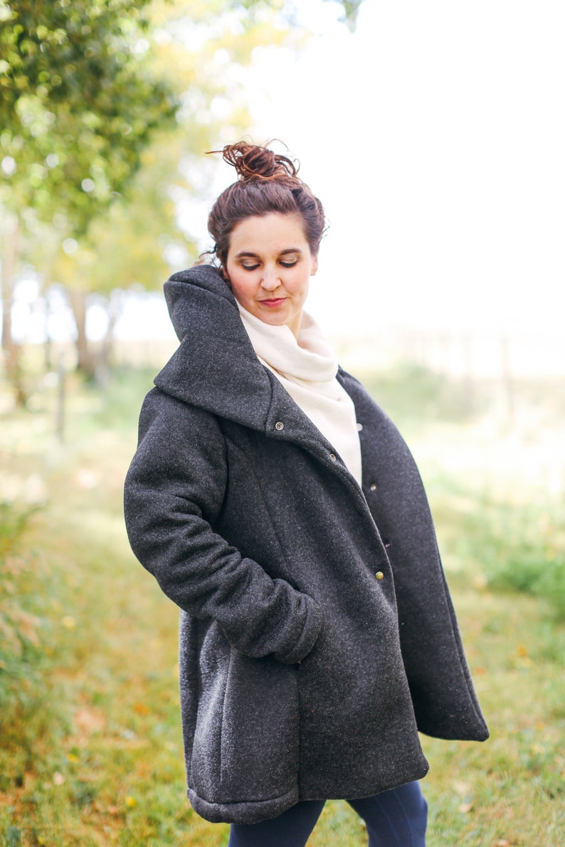 May include: A woman wearing a dark gray, long-sleeved coat with a high collar and a white turtleneck sweater underneath. The coat has a button closure and pockets.