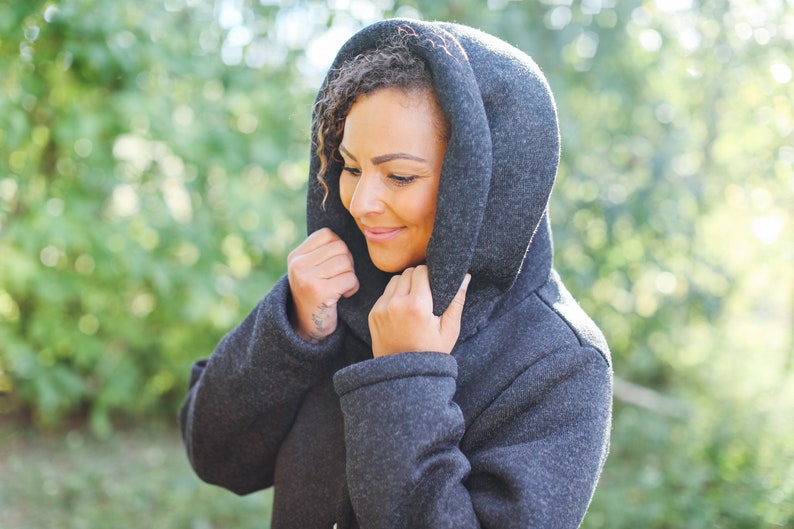 May include: A woman wearing a dark gray hooded jacket with a soft, fuzzy texture. The hood is pulled up around her head, and she is looking down. The background is blurred and out of focus.