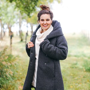 May include: A woman wearing a dark gray hooded coat with a white turtleneck sweater underneath. She is standing in a grassy field with trees in the background.