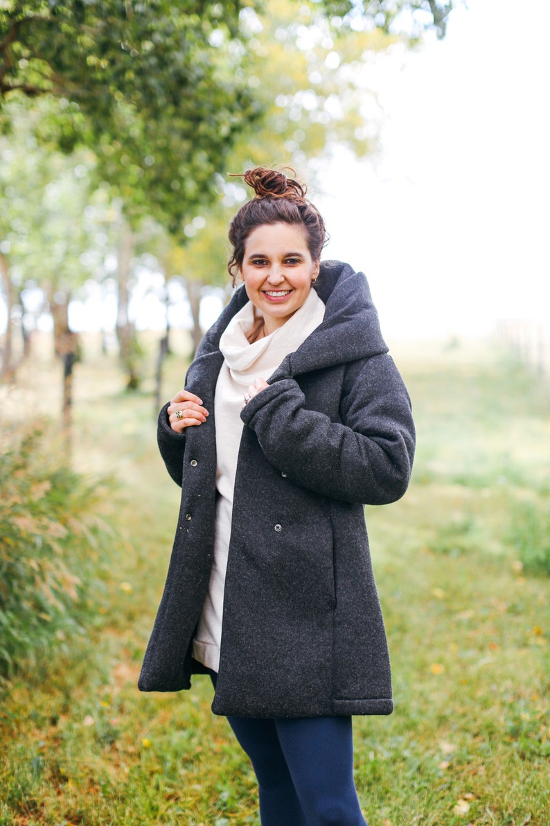 May include: A woman wearing a dark gray hooded coat with a white turtleneck sweater underneath. She is standing in a grassy field with trees in the background.