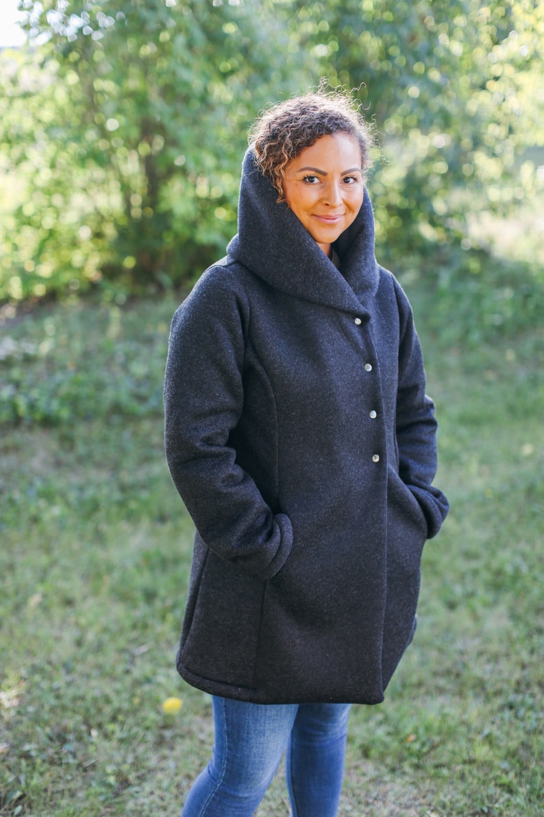May include: A woman wearing a dark gray hooded jacket with a button closure. She is standing in a grassy area with trees in the background.