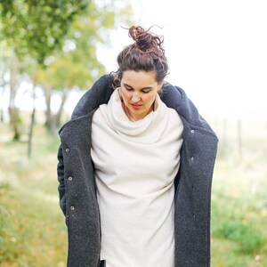 May include: A woman wearing a white turtleneck sweater with a dark gray coat over it. She is wearing dark blue pants and has her hair pulled back in a bun.