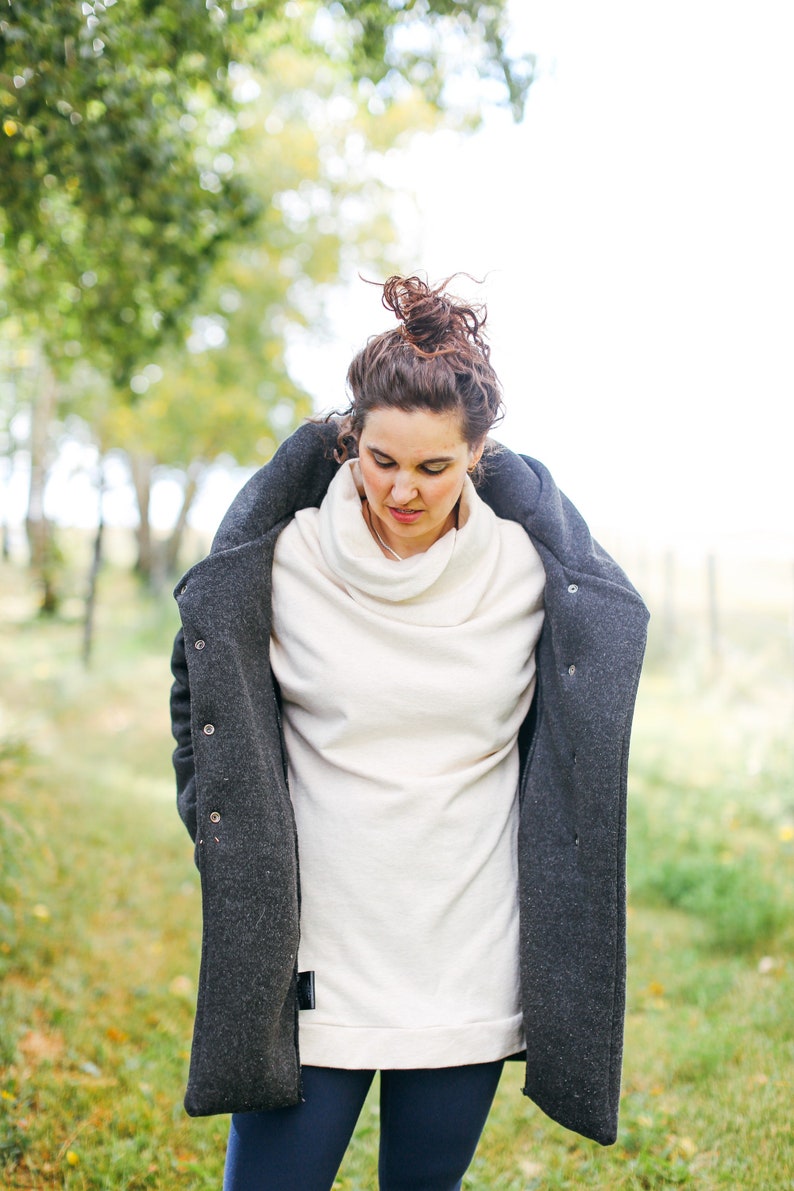 May include: A woman wearing a white turtleneck sweater with a dark gray coat over it. She is wearing dark blue pants and has her hair pulled back in a bun.