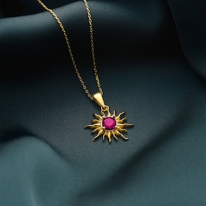 May include: A gold-colored necklace with a sun pendant. The pendant features a central, round, ruby-colored stone surrounded by stylized sun rays. The necklace chain is delicate and gold, set against a dark teal background.