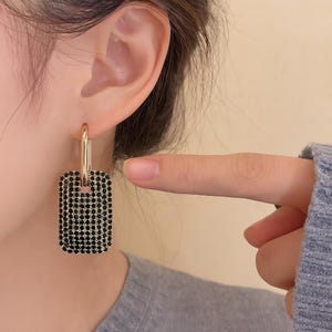 Może przedstawiać: Złote kolczyki koła z prostokątną zawieszką wysadzaną czarnymi kamieniami. Kolczyki są pokazane na uchu, z palcem wskazującym na biżuterię. Prosty i elegancki design.