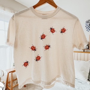 May include: An ivory short-sleeve t-shirt featuring a scattered pattern of small red ladybugs with black spots across the front. The shirt has a crew neckline and is displayed on a wooden hanger. The text COMFORT COLORS is visible in the top right corner.