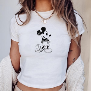May include: White crop top featuring a black and white sketch of Mickey Mouse. The cartoon character is depicted in a walking pose. The shirt has short sleeves and a crew neck.