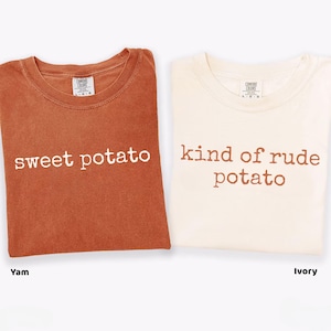May include: Two folded t-shirts, one in a yam colour and the other ivory, are shown. The yam-coloured shirt has the words "sweet potato" printed in white. The ivory shirt has the words "kind of rude potato" printed in brown.