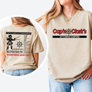 Op de afbeelding: Een beige t-shirt met de rode tekst Cap'n Clark's en de zwarte tekst OTTOMAN EMPIRE op de voorzijde. De achterkant toont een piratengrafiek, een scheepsroer, de tekst Cap'n Clark's Ottoman Empire, een lijst met meubels en de tekst EVERYTHING MUST GO.
