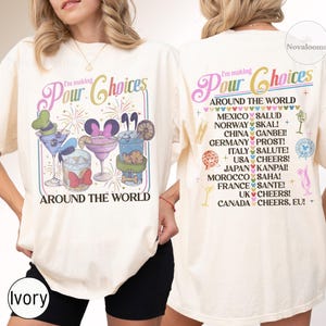 May include: An ivory t-shirt featuring a graphic print on the front and back. The front displays colorful cocktails with character-themed accessories and the text I'm making Pour Choices AROUND THE WORLD. The back lists international toasts.