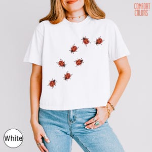 May include: A white short-sleeve t-shirt featuring a graphic print of seven red ladybugs with black spots scattered across the front. The shirt includes the text COMFORT COLORS printed in the upper right corner and a white circular label.