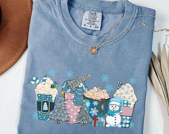 May include: Blue jean colored Comfort Colors t-shirt with a festive winter design. The graphic features decorated coffee cups, Christmas trees, snowflakes, and a snowman. A gold necklace is draped across the shirt. The words "Blue Jean" are at the bottom.