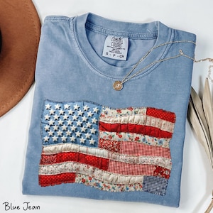 Puede incluir: Camiseta azul vaquero con un diseño de bandera americana cosido. La bandera presenta estrellas blancas sobre un campo azul y rayas rojas y blancas hechas de varias telas estampadas. Un collar dorado con un colgante descansa sobre la camisa.