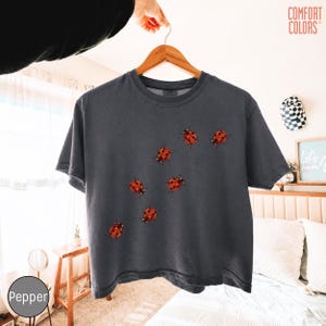 May include: A charcoal gray short-sleeve t-shirt featuring a scattered pattern of red and black ladybug illustrations across the front. The shirt has a crew neckline and is displayed on a wooden hanger against a light-colored background.