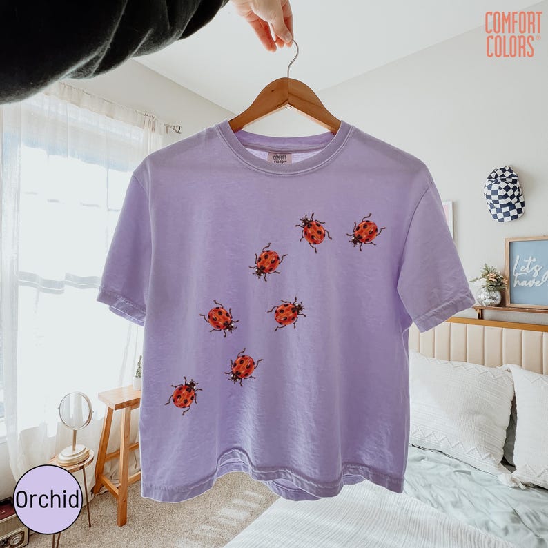 May include: A light purple short-sleeve t-shirt featuring a scattered pattern of red and black ladybug illustrations across the front. The shirt has a crew neckline and a Comfort Colors brand label printed on the inside of the collar.