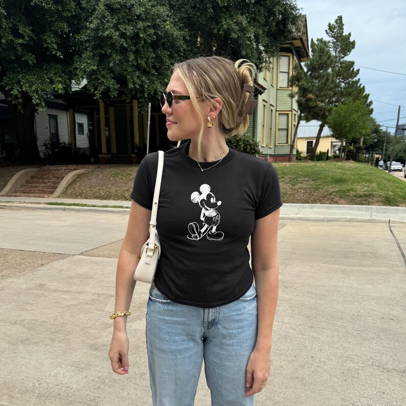 May include: Black t-shirt featuring a white graphic of Mickey Mouse. The shirt is paired with light blue jeans and a white purse. The person is wearing sunglasses and gold jewelry.