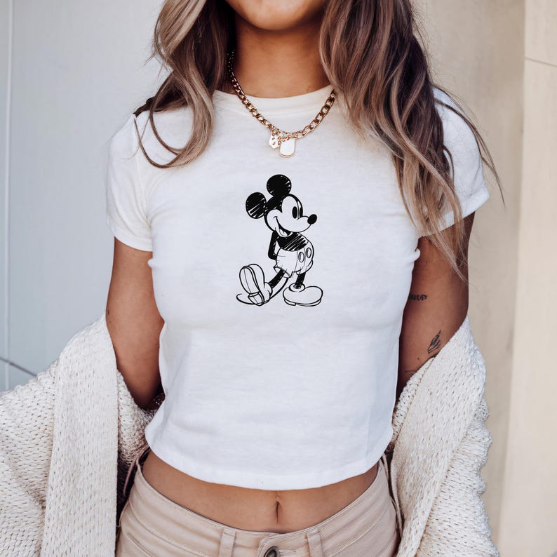 May include: White crop top featuring a black and white sketch of Mickey Mouse. The cartoon character is depicted in a walking pose. The shirt has short sleeves and a crew neck.