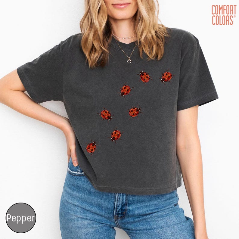 May include: A charcoal gray short-sleeve t-shirt featuring a scattered pattern of small red ladybugs with black spots across the front. The shirt has a crew neckline and the text COMFORT COLORS is printed in the upper right corner of the image.