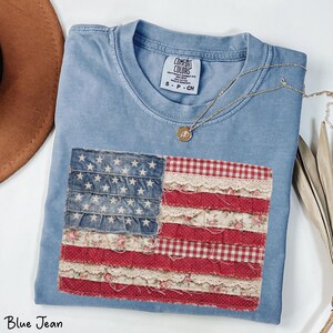 Puede incluir: Una camiseta azul Comfort Colors con un diseño de bandera americana de patchwork. La bandera presenta estrellas de mezclilla, rayas florales y de cuadros rojos y blancos, y detalles de encaje. Un collar dorado con un colgante descansa sobre la camisa.